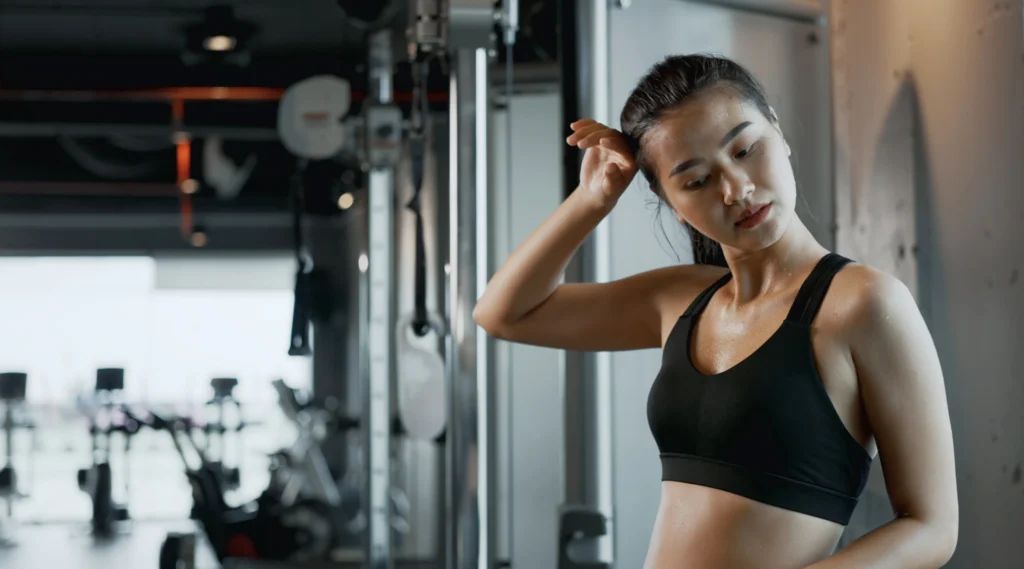 Perempuan berkeringat di gym setelah workout, mengenakan sports bra hitam. Gambar ini menggambarkan pentingnya menjaga area kewanitaan tetap bersih dan kering setelah berolahraga untuk mencegah infeksi dan iritasi.