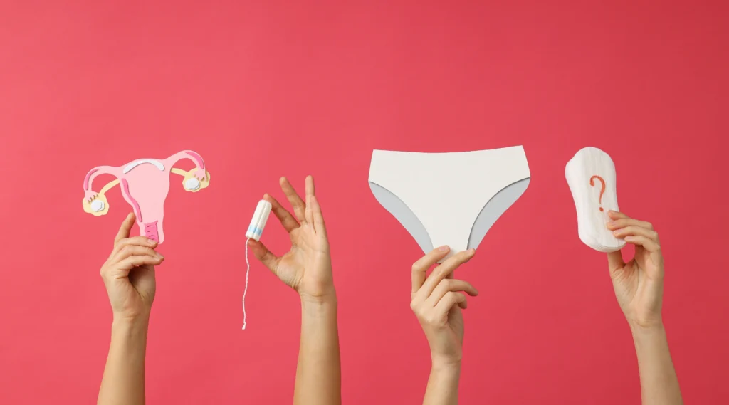 Tangan-tangan perempuan memegang ilustrasi rahim, tampon, celana dalam, dan pembalut dengan latar belakang merah muda, menggambarkan pentingnya perawatan area kewanitaan saat haid
